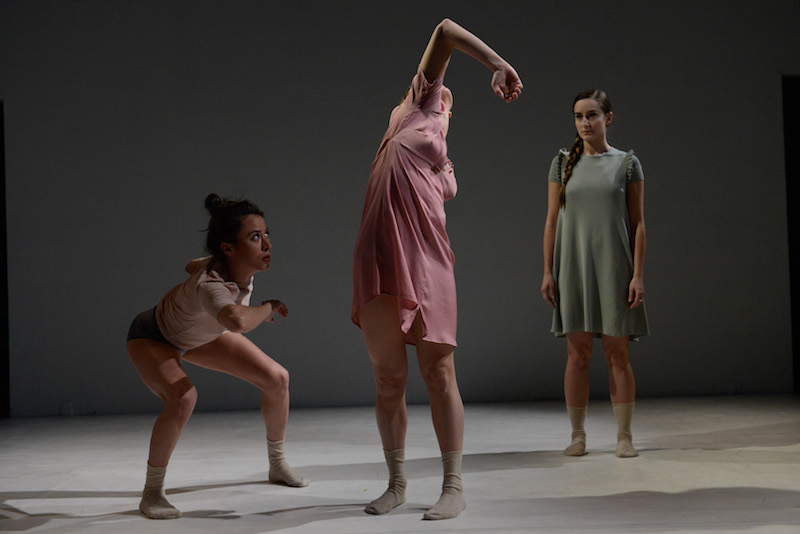 Three women in socked feet. One is crouching down and looking up at the center figure whose elbow is pointing up. Another dancer in the background watches.